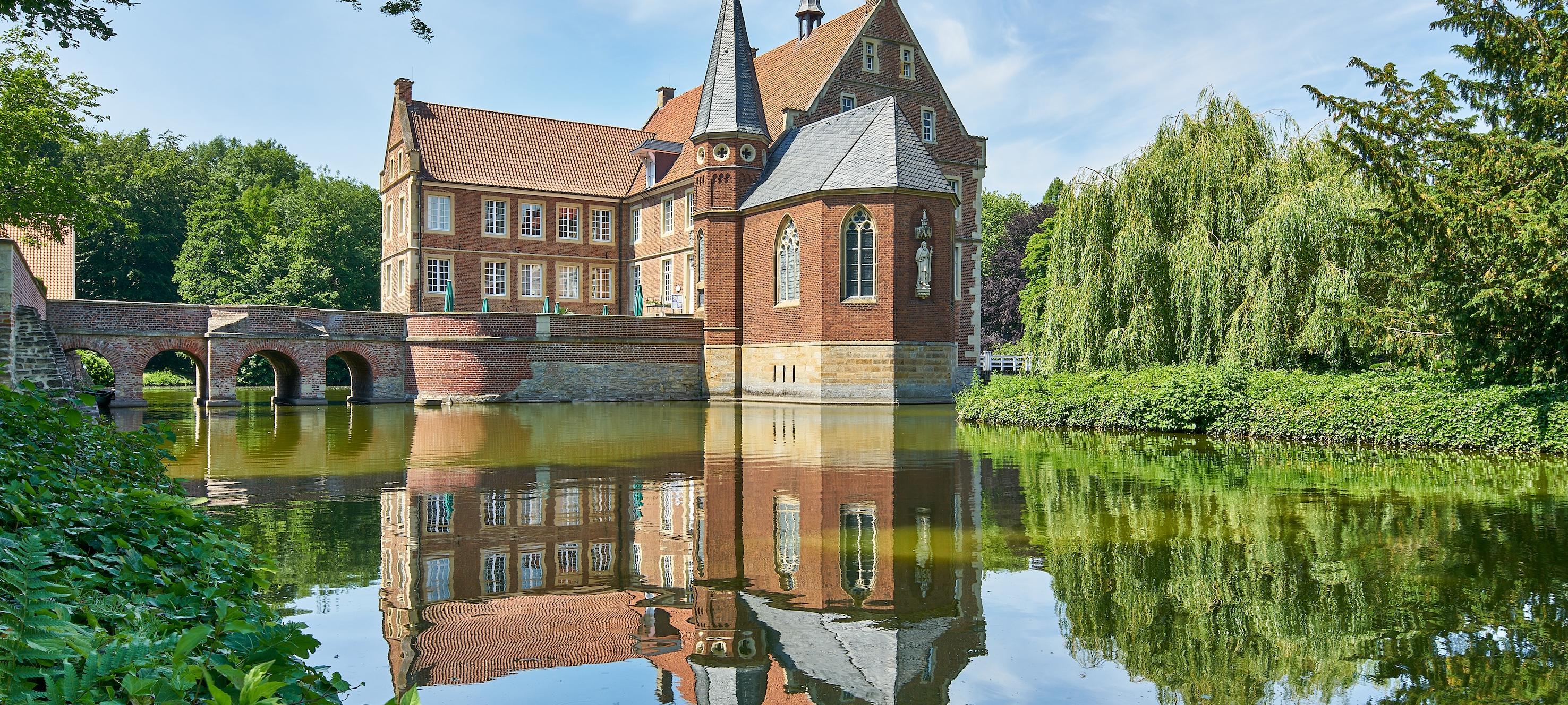 Eine kleine Burg vor einem See im Sonnenschein.